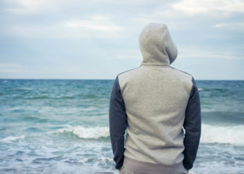 Man stands on the beach in a tracksuit with a hood and looking a A man stands on the beach in a tracksuit with a hood and looking at the sea. Cloudy weather, rear view.